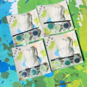 May include: Four paint-your-own frog figurines with green paint pots and brushes. Each frog is sitting on a green lily pad with green leaves and a blue pond. The background is a white paper with green frog footprints.