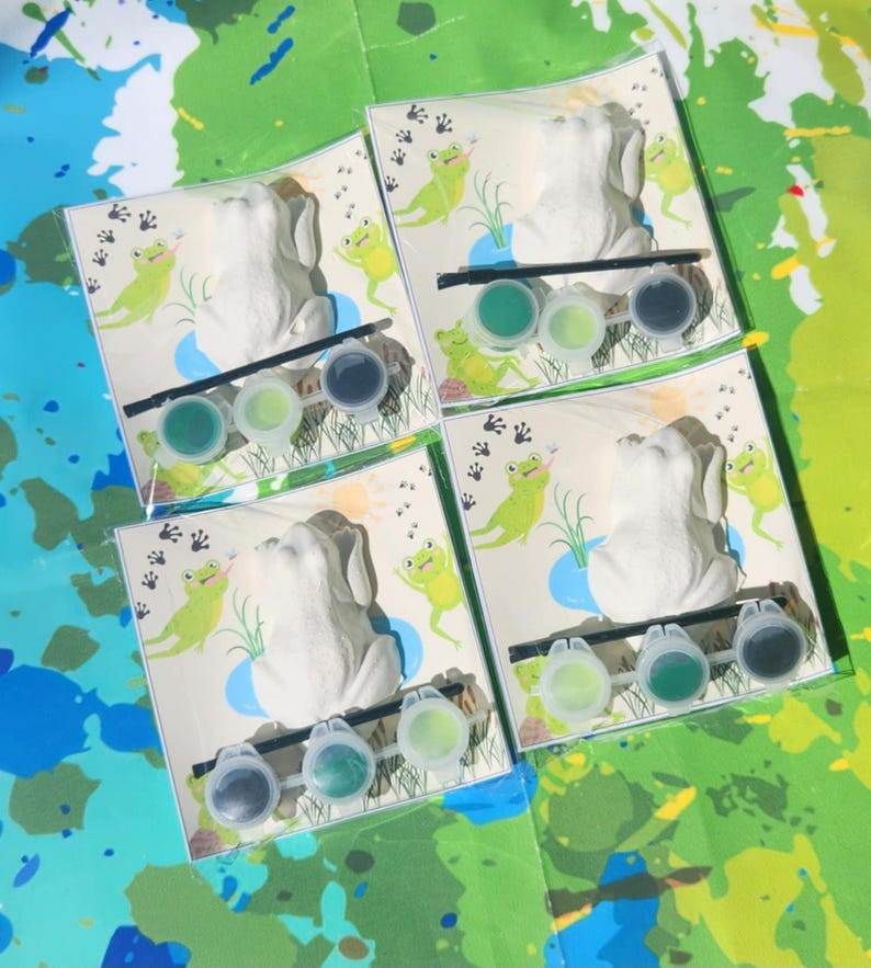 May include: Four paint-your-own frog figurines with green paint pots and brushes. Each frog is sitting on a green lily pad with green leaves and a blue pond. The background is a white paper with green frog footprints.