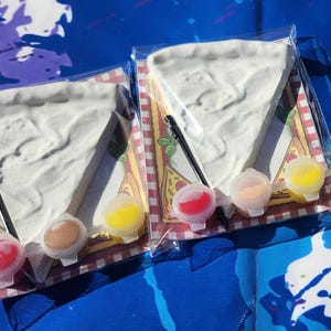 May include: Three paint-your-own pizza slice kits. Each kit includes a white ceramic pizza slice, a small paintbrush, and three small containers of paint: red, yellow, and brown.