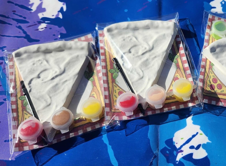 May include: Three paint-your-own pizza slice kits. Each kit includes a white ceramic pizza slice, a small paintbrush, and three small containers of paint: red, yellow, and brown.