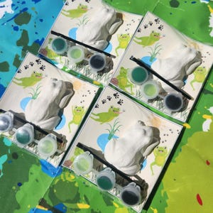 May include: Four paint-your-own frog figurines with green, blue, and black paint pots and brushes. Each frog is on a card with a green background and a pond scene.