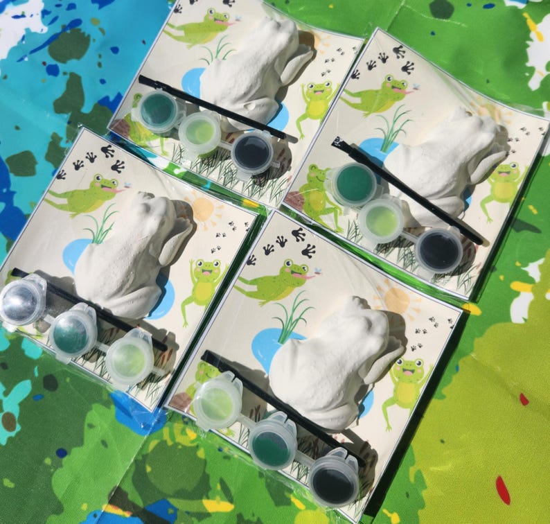May include: Four paint-your-own frog figurines with green, blue, and black paint pots and brushes. Each frog is on a card with a green background and a pond scene.