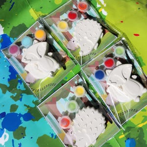May include: Four paint-your-own ceramic animal kits. Each kit includes a ceramic animal, a paintbrush, and a set of paint pots. The animals are a fox, a hedgehog, and two hedgehogs. The paint pots are in various colors, including red, blue, green, yellow, and orange.