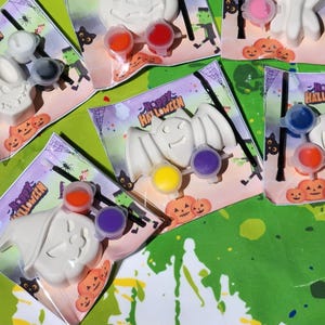May include: A set of five Halloween-themed paint-your-own kits. Each kit includes a white ceramic figurine, a small paintbrush, and three small containers of paint. The figurines include a skull, a bat, a pumpkin, and two ghosts. The packaging is decorated with the words "Happy Halloween".