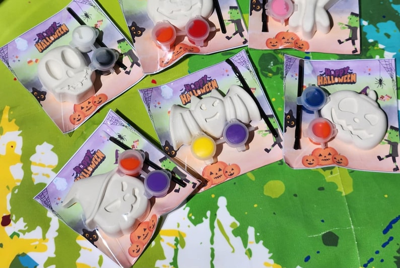 May include: A set of five Halloween-themed paint-your-own kits. Each kit includes a white ceramic figurine, a small paintbrush, and three small containers of paint. The figurines include a skull, a bat, a pumpkin, and two ghosts. The packaging is decorated with the words "Happy Halloween".