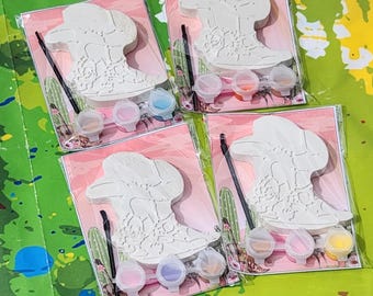 May include: Four paint-your-own ceramic cowboy boot and hat kits. Each kit includes a white ceramic boot and hat, a small paintbrush, and four small bottles of paint in various colours.