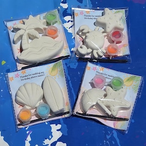 Puede incluir: Cuatro kits de manualidades con temática de playa en envases transparentes. Cada kit incluye una figura de yeso, botes de pintura y un pincel. Las figuras incluyen una palmera, un cangrejo, una concha y una estrella de mar. El texto en la tarjeta dice "Thanks for making my birthday shore-tastic!"