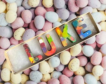 May include: A wooden box filled with colorful crayon letters spelling out the name "LUKE" surrounded by pastel-colored mini chocolate eggs.