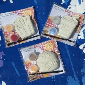 Puede incluir: Tres kits de pintura para hacer uno mismo con formas de comida rápida: patatas fritas, un vaso de cola y una hamburguesa. Cada kit incluye botes de pintura y un pincel. El embalaje dice "Thanks for making party Buns of Fun!"