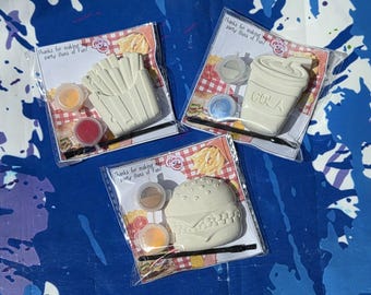 May include: Three paint-your-own kits featuring fast food shapes: french fries, a cola cup, and a hamburger. Each kit includes paint pots and a paintbrush. The packaging reads "Thanks for making party Buns of Fun!"