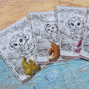 May include: A set of clear plastic bags containing colouring cards featuring a cartoon monkey, a treasure chest, and a sun, each paired with a small banana-shaped crayon in colours including olive green, light brown, pink, and dark blue.
