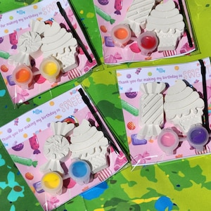 May include: Three sets of DIY paint your own candy and cupcake shaped ornaments. Each set includes two ornaments, paint, and a paintbrush. The packaging has a pink background with a candy theme and the text "Thank you for making my birthday so sweet".