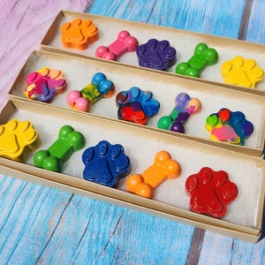 May include: A set of colorful crayons shaped like paw prints and bones, packaged in three brown boxes. The crayons are arranged in a row, with the paw prints on the left and the bones on the right.