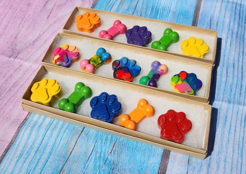 May include: A set of colorful crayons shaped like paw prints and bones, packaged in three brown boxes. The crayons are arranged in a row, with the paw prints on the left and the bones on the right.