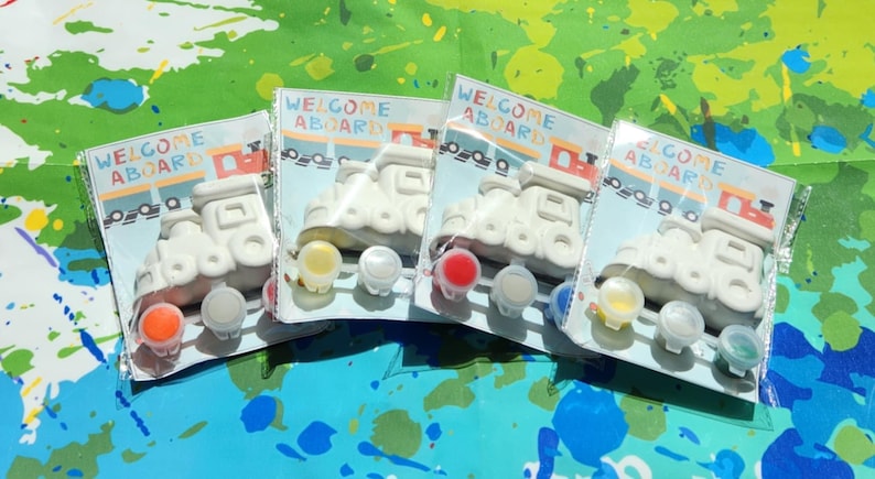 May include: Four white ceramic train engine figurines with the words "Welcome Aboard" on the front. Each figurine comes with four small pots of paint in different colors: red, yellow, white, and green.