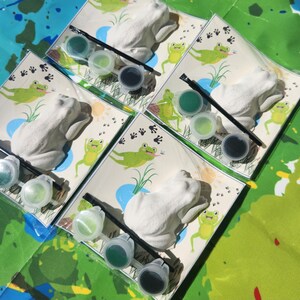 May include: Four sets of frog-shaped ceramic figurines with paint and brushes for painting. Each set includes three paint pots, a brush, and a frog figurine.