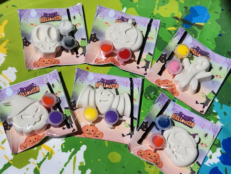 May include: Set of six Halloween-themed paint-your-own ceramic ornaments. Each ornament features a different Halloween character, including a ghost, pumpkin, bat, witch, and two skulls. Each ornament comes with a small paintbrush and three paint pots.