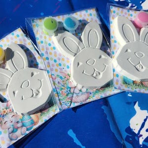 May include: Three Easter bunny paint-your-own kits with colorful paint pots. Each kit includes a white ceramic bunny figurine, a paintbrush, and three small paint pots in different colors.