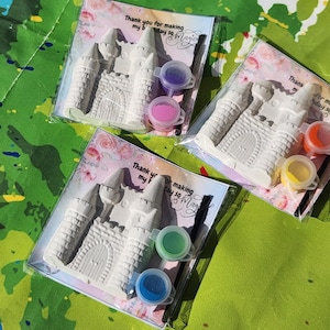 May include: Three sets of DIY castle painting kits. Each kit includes a white castle figurine, two paint pots, and a small paintbrush, all packaged in a clear plastic bag. The text on the card reads "Thank you for making my day so Magic."