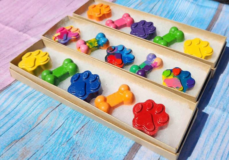 May include: Three boxes filled with colorful crayon paw prints and bone shapes. The crayons are arranged in rows and are ready to be used for coloring.