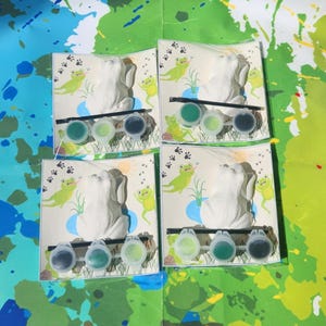 May include: Four paint-your-own frog figurines with green paint pots and brushes. Each frog is sitting on a green lily pad with green frog footprints in the background.