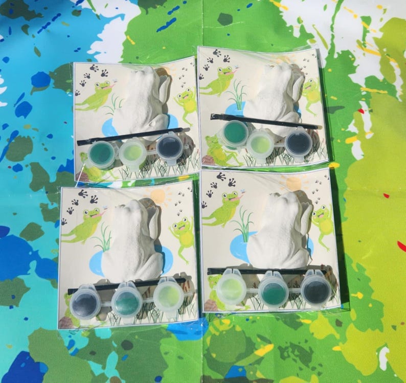 May include: Four paint-your-own frog figurines with green paint pots and brushes. Each frog is sitting on a green lily pad with green frog footprints in the background.