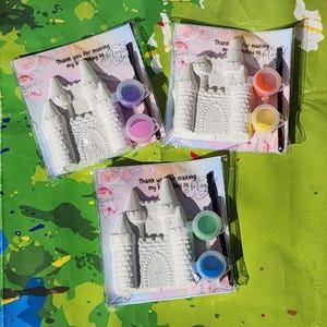 May include: Three paint-your-own castle kits. Each kit includes a white castle figurine, paint pots in various colors, and a paintbrush. The packaging features text that reads "Thank you for making my bday so Magic."