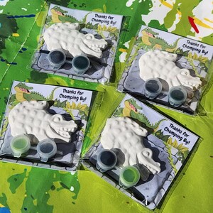 Może przedstawiać: Cztery przezroczyste plastikowe torebki, z kt&oacute;rych każda zawiera białą figurkę gipsową w kształcie aligatora, dwa małe pojemniki z farbą i kartkę z napisem "Thanks for Chomping By!". Tło to zielono-niebieska pomalowana powierzchnia.