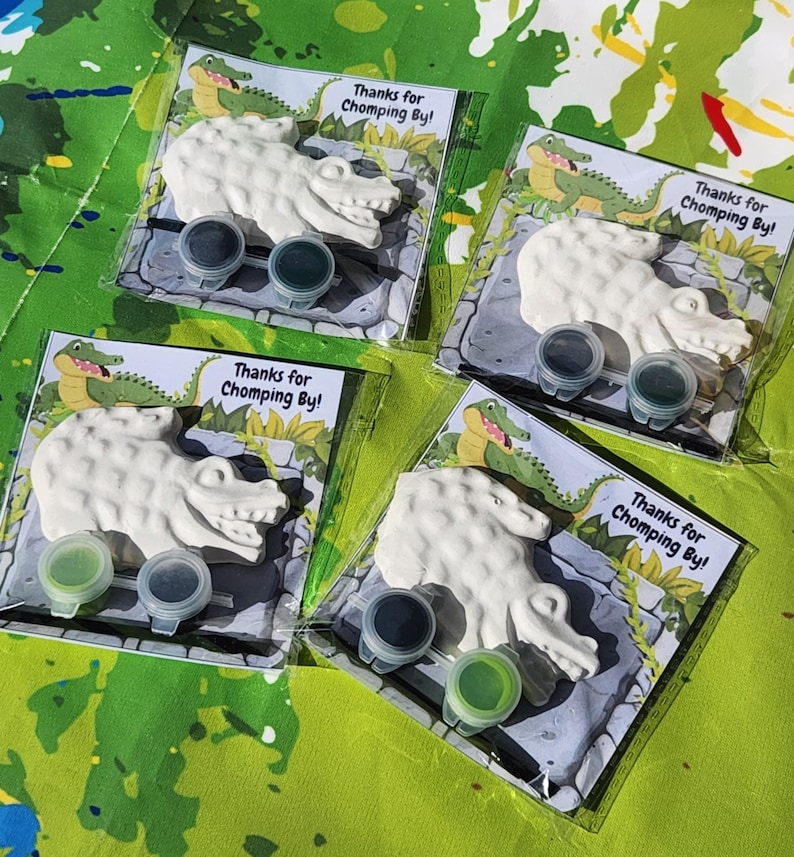 Może przedstawiać: Cztery przezroczyste plastikowe torebki, z kt&oacute;rych każda zawiera białą figurkę gipsową w kształcie aligatora, dwa małe pojemniki z farbą i kartkę z napisem "Thanks for Chomping By!". Tło to zielono-niebieska pomalowana powierzchnia.