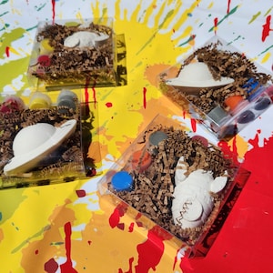 May include: Four clear plastic boxes filled with brown packing peanuts and small white ceramic figurines. Each box contains a different figurine, including a spaceship, a rocket, and a pig. The boxes also contain small bottles of paint.