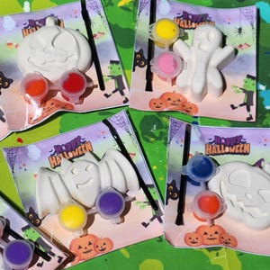 May include: Four Halloween-themed paint-your-own kits with a ghost, bat, pumpkin, and skull. Each kit includes a white ceramic figure, paint pots, and a paintbrush. The kits are packaged in clear plastic bags with a colorful Halloween-themed design.