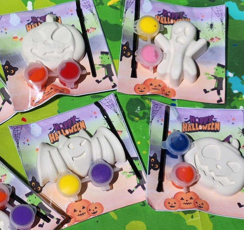 May include: Four Halloween-themed paint-your-own kits with a ghost, bat, pumpkin, and skull. Each kit includes a white ceramic figure, paint pots, and a paintbrush. The kits are packaged in clear plastic bags with a colorful Halloween-themed design.