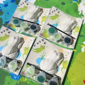 May include: Four sets of frog-shaped ceramic figurines with paint pots and brushes for painting. Each set includes a frog figurine, three paint pots, and a paintbrush. The paint pots are green, light green, and dark blue.
