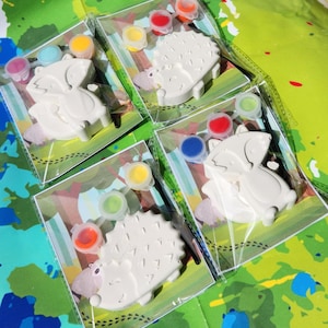 May include: Four paint-your-own animal figurines packaged in clear plastic bags. Each bag contains a white ceramic figurine, a paintbrush, and four small pots of paint. The figurines are a raccoon, a hedgehog, and two hedgehogs. The paint colors are red, blue, green, yellow, and orange.