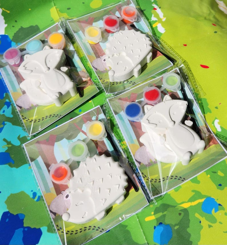 May include: Four paint-your-own animal figurines packaged in clear plastic bags. Each bag contains a white ceramic figurine, a paintbrush, and four small pots of paint. The figurines are a raccoon, a hedgehog, and two hedgehogs. The paint colors are red, blue, green, yellow, and orange.
