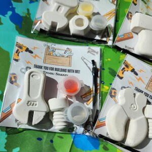 May include: A DIY craft kit for children. The kit includes a white ceramic wrench, a white ceramic pipe, a small paintbrush, a small bottle of red paint, a small bottle of white paint, and a small bottle of yellow paint. The kit comes with a card that says "Thank you for building with me! From: Snazzy".