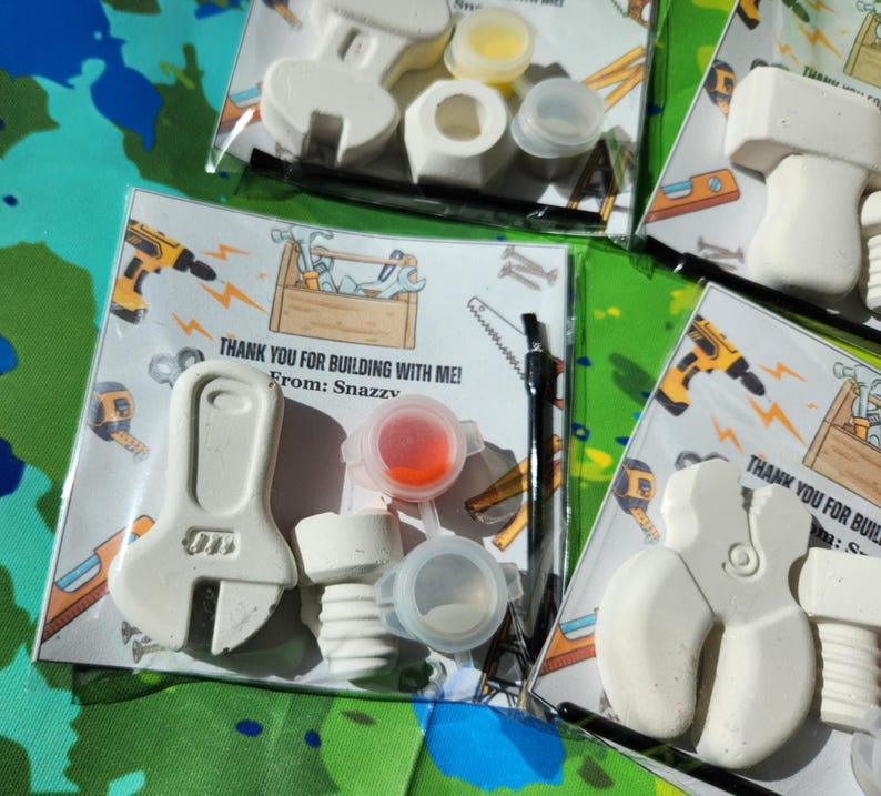 May include: A DIY craft kit for children. The kit includes a white ceramic wrench, a white ceramic pipe, a small paintbrush, a small bottle of red paint, a small bottle of white paint, and a small bottle of yellow paint. The kit comes with a card that says "Thank you for building with me! From: Snazzy".