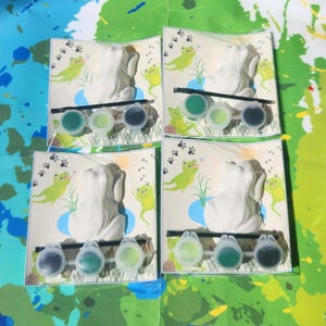 May include: Four paint-your-own frog figurines with paint pots and brushes. Each frog is sitting on a green lily pad with a blue pond behind it. The background is a green and blue watercolor pattern.