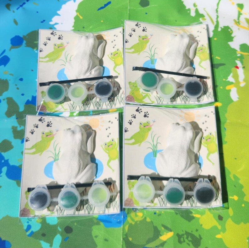 May include: Four paint-your-own frog figurines with paint pots and brushes. Each frog is sitting on a green lily pad with a blue pond behind it. The background is a green and blue watercolor pattern.