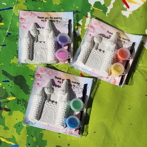 May include: Three packaged craft kits featuring a white castle, paint pots, and a paintbrush. Each kit includes a card with the text "Thank you for making my bday so Magic". The background is a green surface with colorful paint splatters.