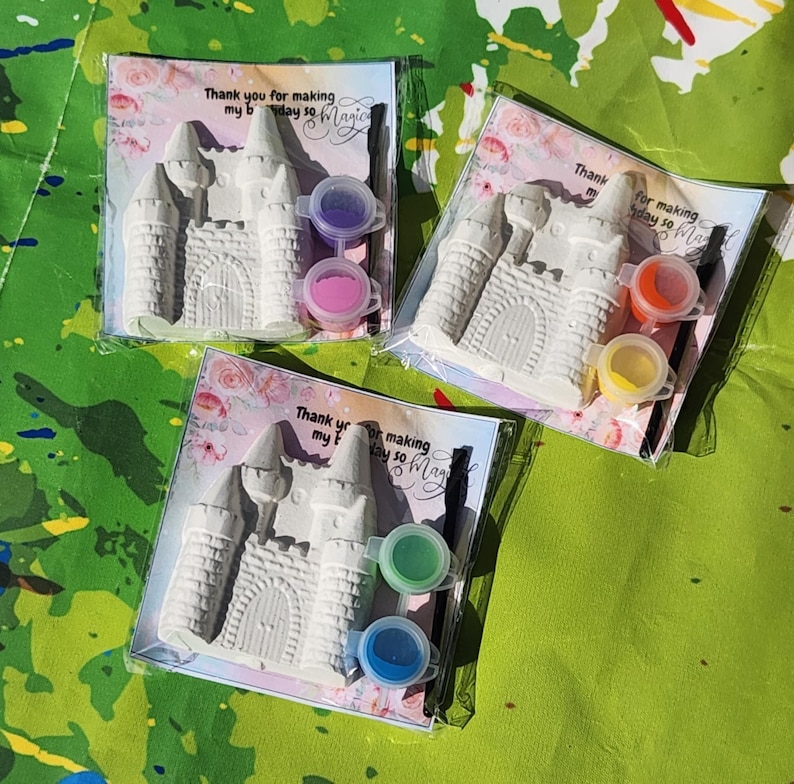 May include: Three packaged craft kits featuring a white castle, paint pots, and a paintbrush. Each kit includes a card with the text "Thank you for making my bday so Magic". The background is a green surface with colorful paint splatters.