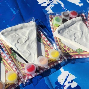 May include: A set of four paint-your-own pizza slices with a variety of paint colors. The pizza slices are made of a white ceramic material and are ready to be painted. The paint colors are yellow, orange, red, and green. The pizza slices are on a white background with a red and white checkered pattern.