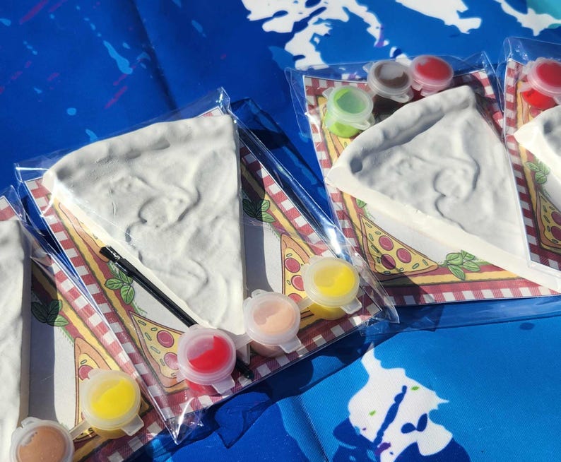 May include: A set of four paint-your-own pizza slices with a variety of paint colors. The pizza slices are made of a white ceramic material and are ready to be painted. The paint colors are yellow, orange, red, and green. The pizza slices are on a white background with a red and white checkered pattern.
