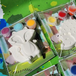 May include: Two paint-your-own ceramic animal figurines, a fox and a hedgehog, with paint pots in various colors. The fox is on a green background with brown footprints, and the hedgehog is on a green background with brown footprints.