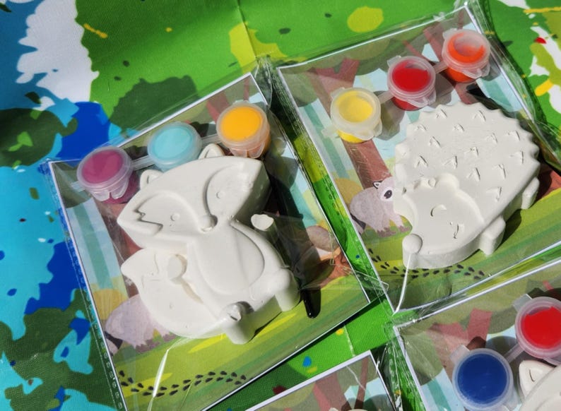 May include: Two paint-your-own ceramic animal figurines, a fox and a hedgehog, with paint pots in various colors. The fox is on a green background with brown footprints, and the hedgehog is on a green background with brown footprints.