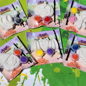 May include: Six Halloween-themed paint-your-own kits with white ceramic figures of a ghost, bat, pumpkin, skull, and witch. Each kit includes two small paint pots and a paintbrush. The kits are packaged on white cardstock with the words "Happy Halloween" printed on them.