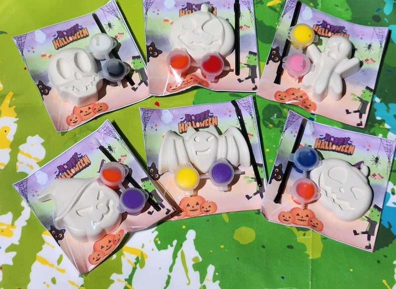 May include: Six Halloween-themed paint-your-own kits with white ceramic figures of a ghost, bat, pumpkin, skull, and witch. Each kit includes two small paint pots and a paintbrush. The kits are packaged on white cardstock with the words "Happy Halloween" printed on them.