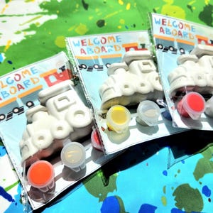 May include: Three paint-your-own ceramic train engine kits with the words "Welcome Aboard" on the packaging. Each kit includes a white ceramic train engine, red, yellow, and blue paint pots, and a paintbrush.