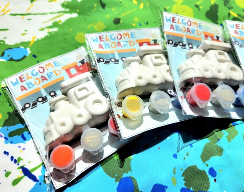May include: Three paint-your-own ceramic train engine kits with the words "Welcome Aboard" on the packaging. Each kit includes a white ceramic train engine, red, yellow, and blue paint pots, and a paintbrush.