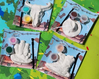 May include: Four paint-your-own ceramic kits with different western themes. Each kit includes a ceramic piece, paint pots, and a paintbrush. The themes are a skull, a cowboy hat, a cactus, and a cowboy boot.